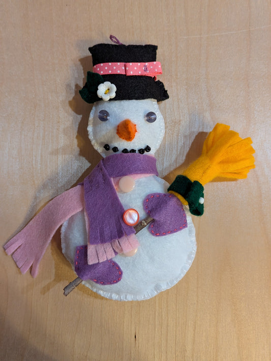 Front view of a handmade snowman ornament with a black hat, purple and pink scarves, and a yellow broom. The snowman ornament is hand stitched with hand applique featuring bead and button accents.