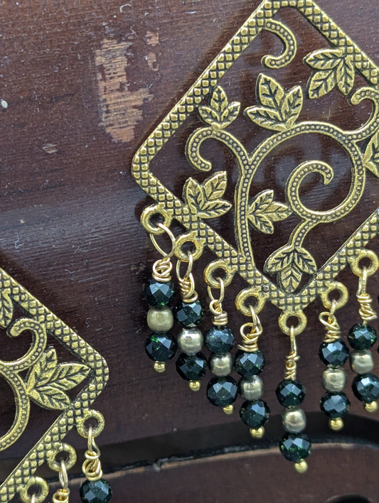 Close up of a set of earrings decorated with golden vines and richly luminous green Swarovski crystals and gold tone pearl seed beads