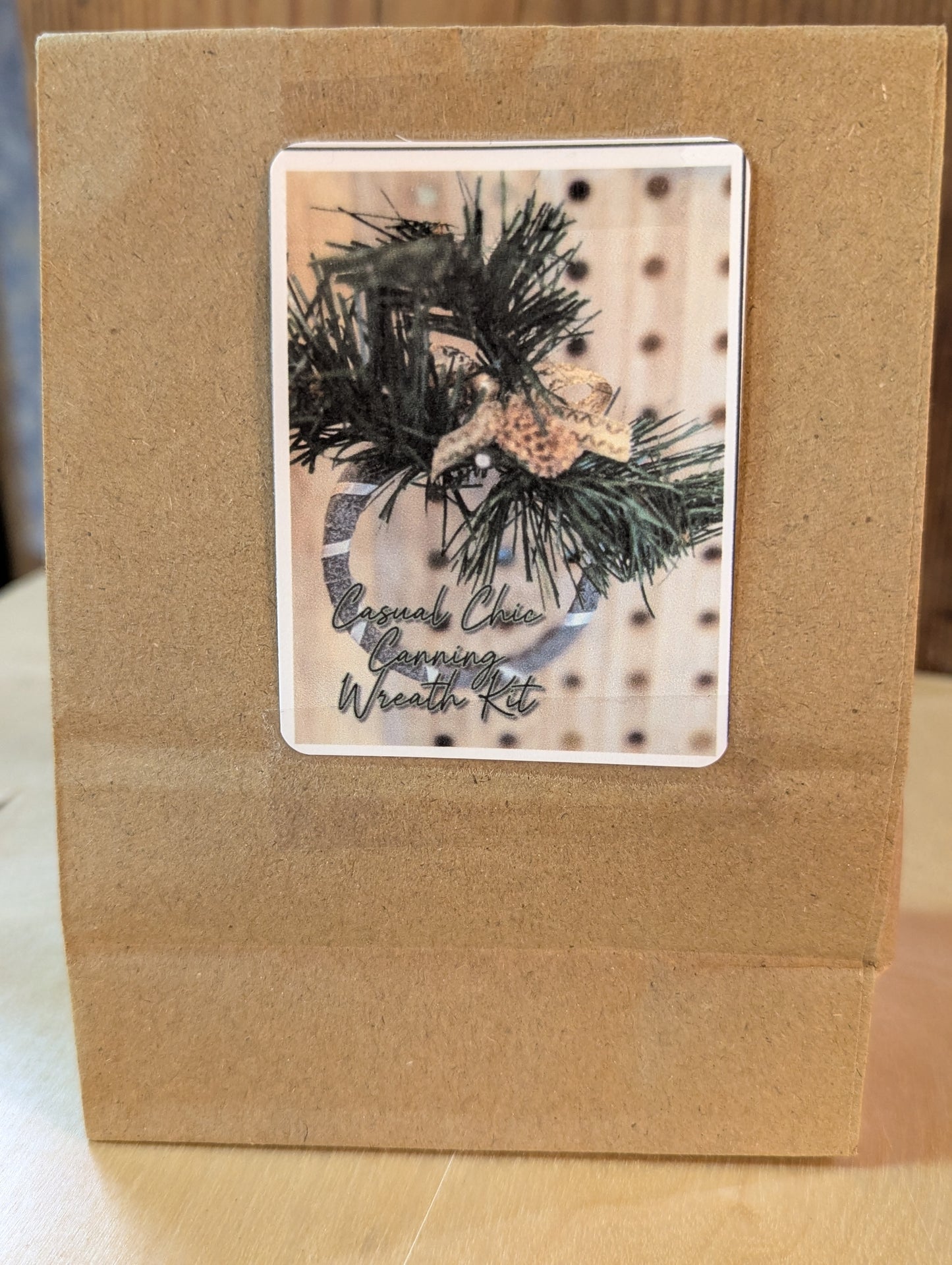 Kraft paper bag with a decorative label featuring the do it yourself canning ring wreath ornament kit in the casual chic style, grey and white with gold ornaments and faux pine branches.