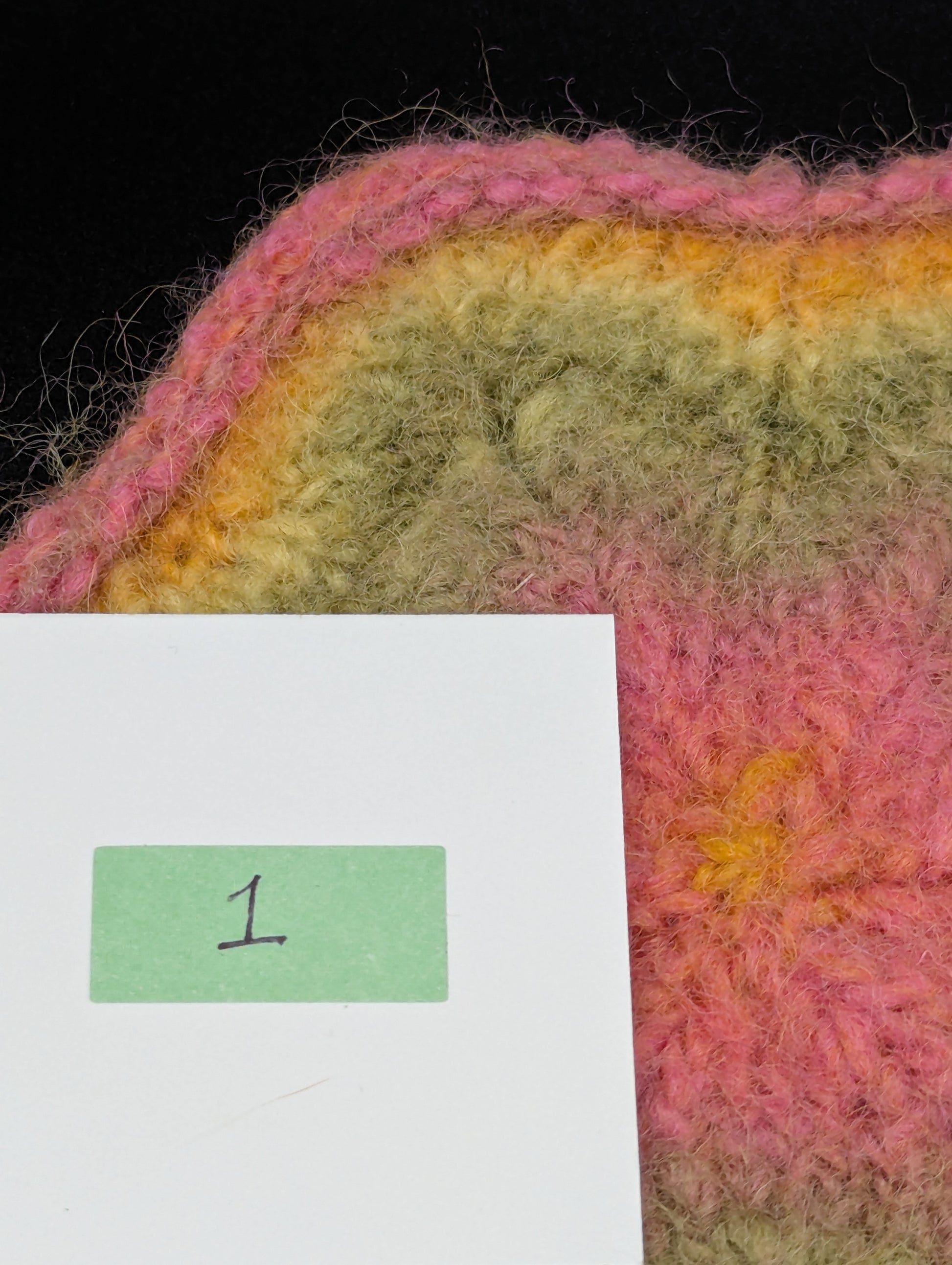 Close up of a multicolored knitted and felted coaster in shades of pink, green and yellow. This coaster is style number 1. 