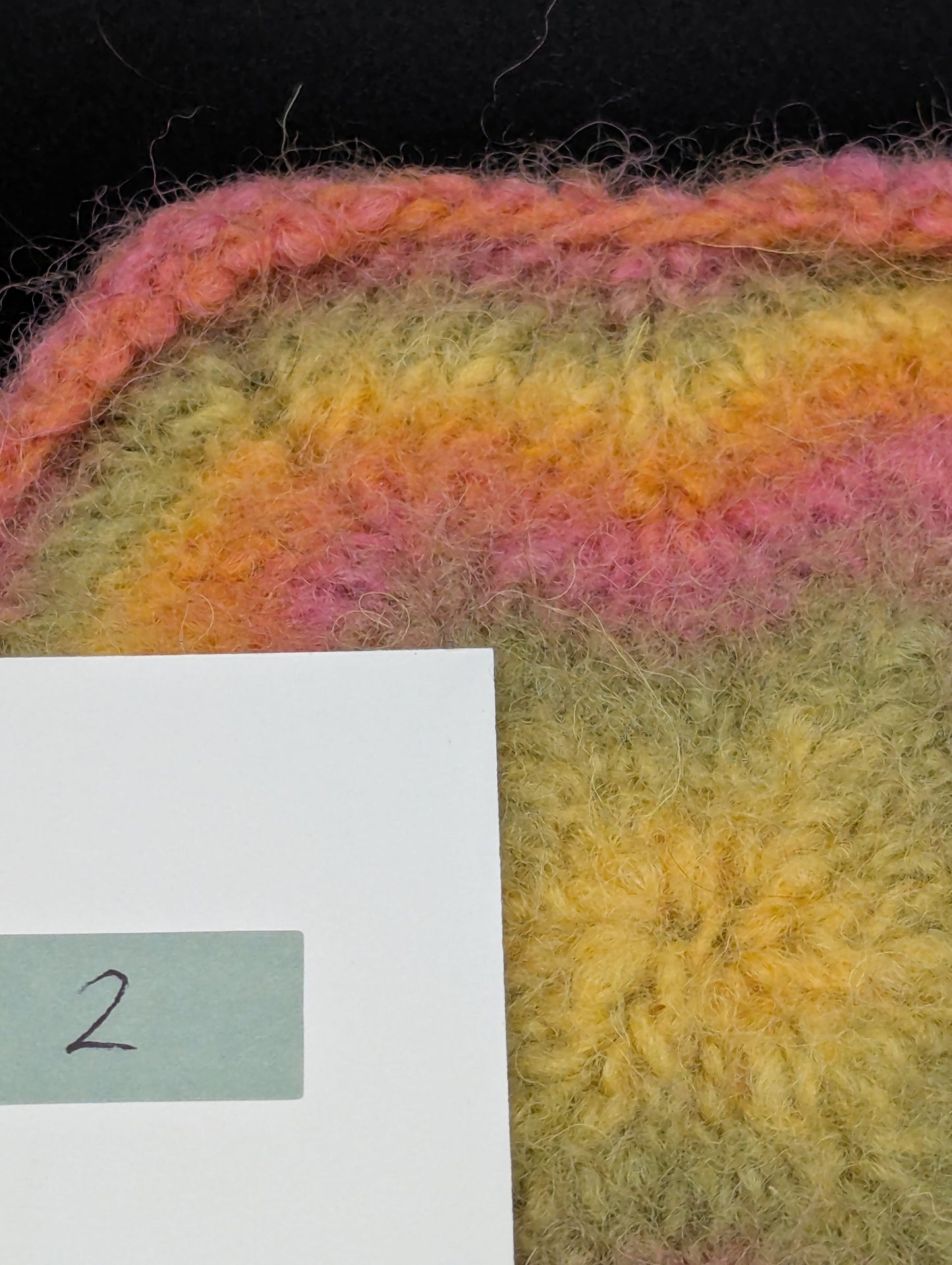 Close up of a multicolored knitted and felted coaster in shades of pink, green and yellow. This coaster is style number 2. 
