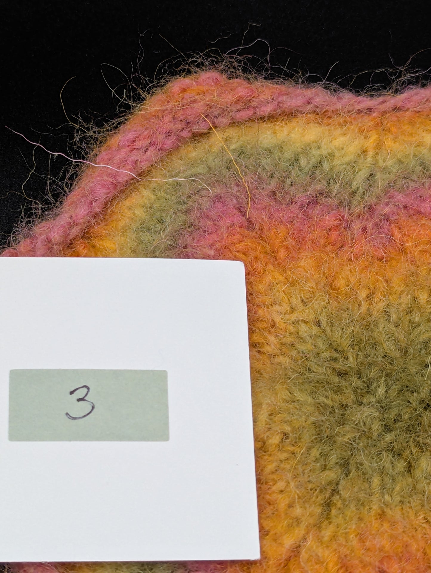 Close up of a multicolored knitted and felted coaster in shades of pink, green and yellow. This coaster is style number 3. 