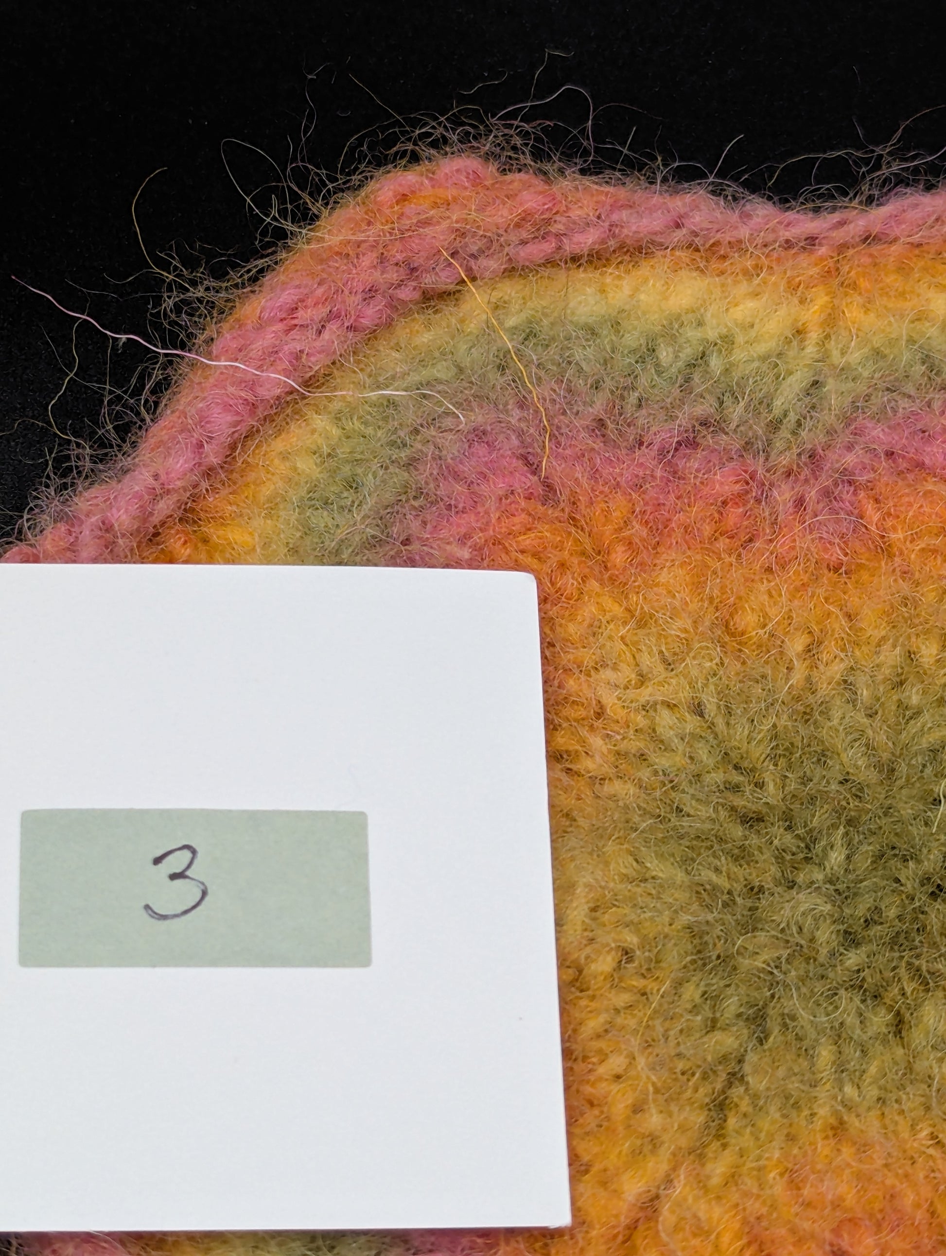 Close up of a multicolored knitted and felted coaster in shades of pink, green and yellow. This coaster is style number 3. 