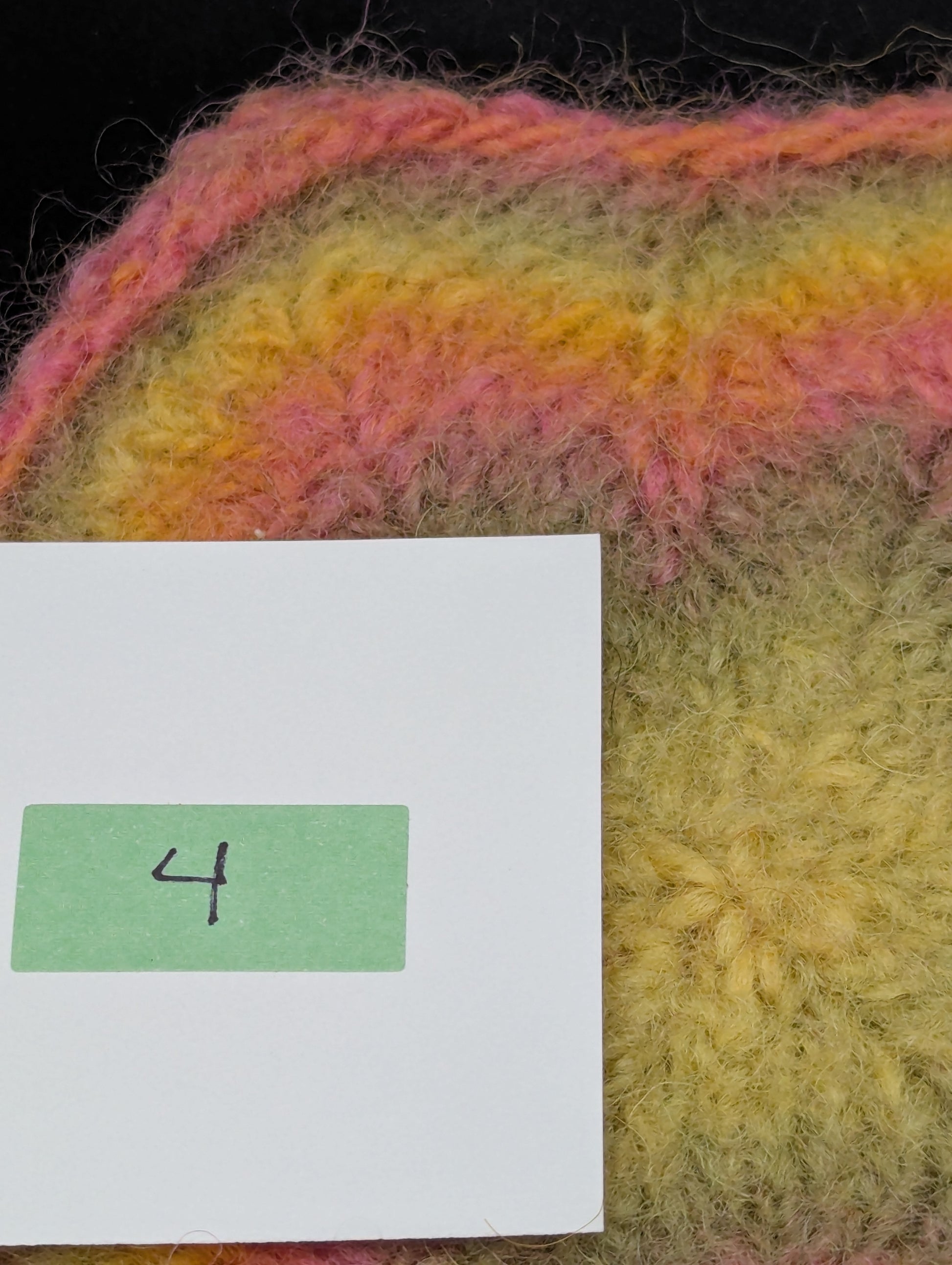 Close up of a multicolored knitted and felted coaster in shades of pink, green and yellow. This coaster is style number 4. 