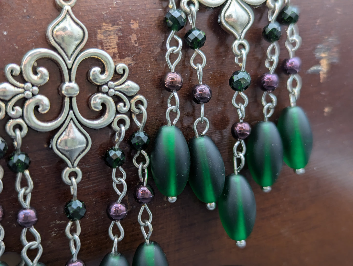 Close up of earrings featuring silver fleur-de-lis with green glass beads highlighted by green crystal beads and Garnet gemstones.