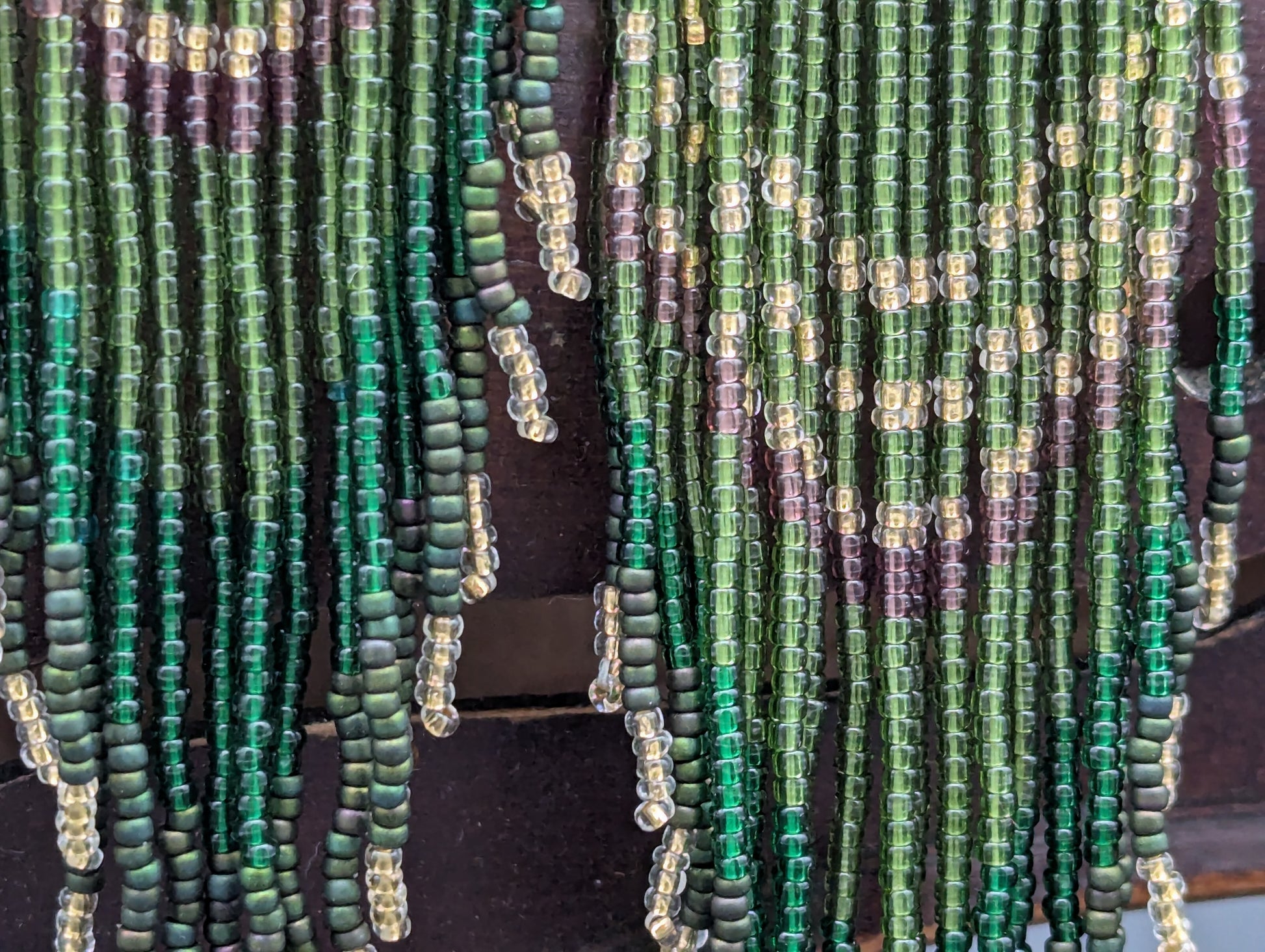 Close up of a pair of hand beaded earrings in green, gold, clear, purple, turquoise and silver glass seed beads trail down from a gold teardrop.
