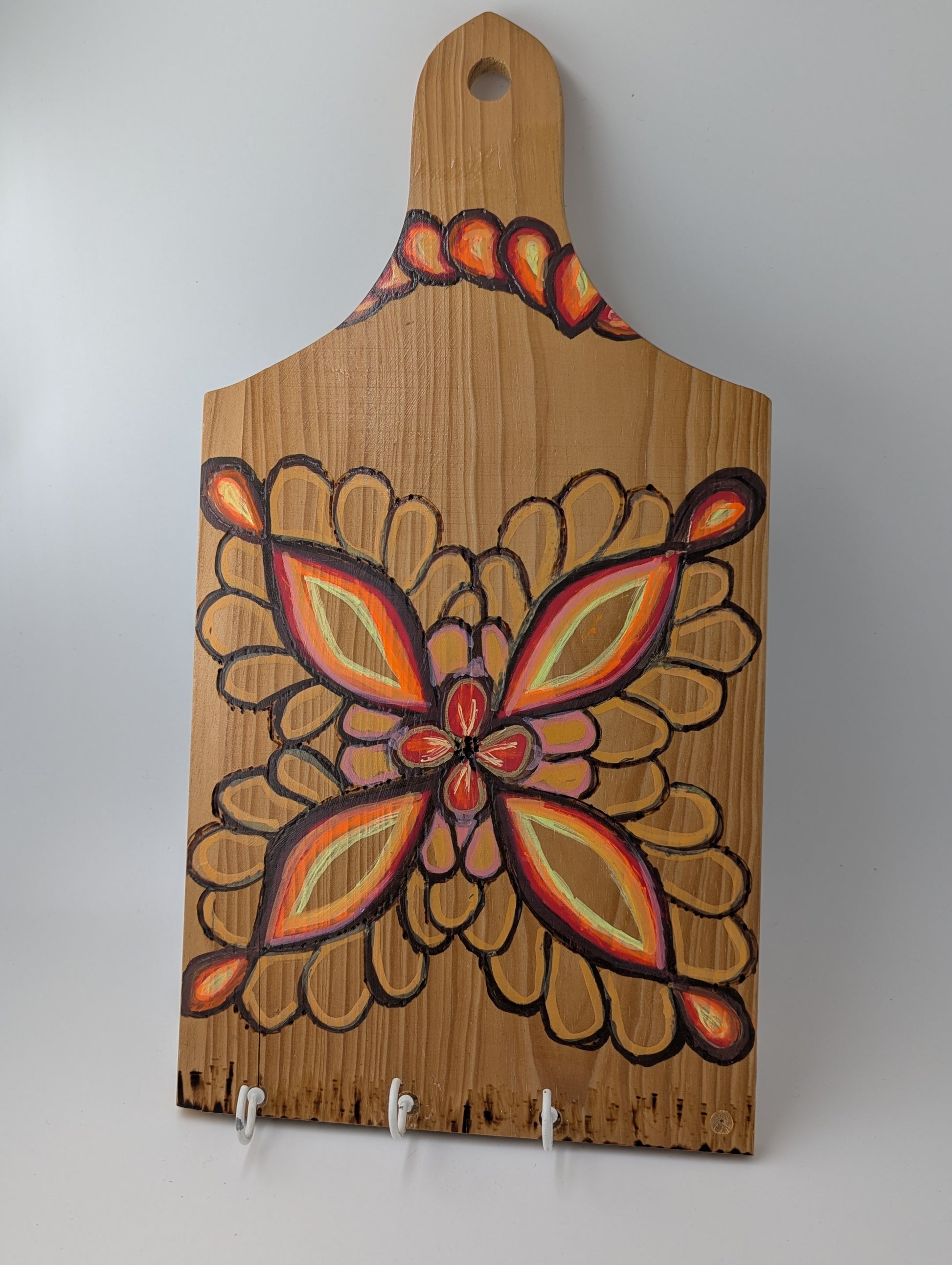 Decorative wooden cutting board with a wood burned and tole painted flower design and 3 cup hooks along the lower edge.