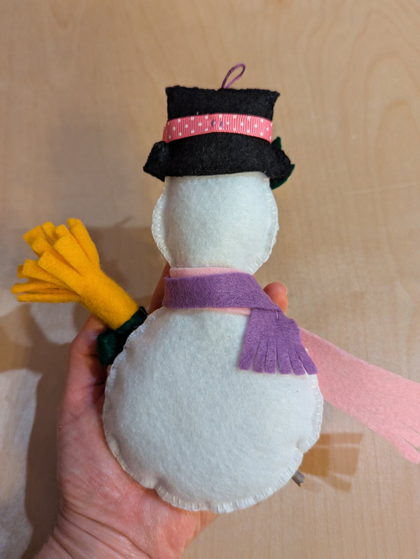 Back view of a handmade snowman ornament with a black hat, purple and pink scarves, and a yellow broom. The snowman ornament is hand stitched with hand applique.