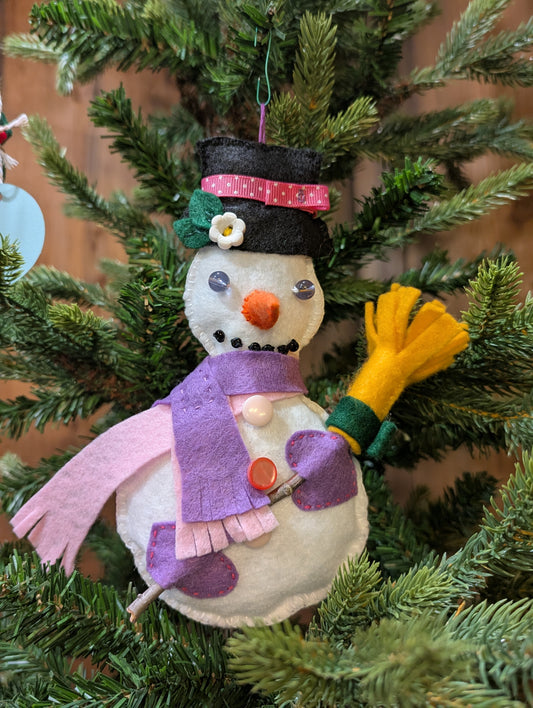 Decorative snowman ornament with pink and lavender scarves and lavender gloves. The snowman ornament is one of a kind, hand stitched, featuring hand applique and button and bead accents. Shown hanging on a Christmas tree.