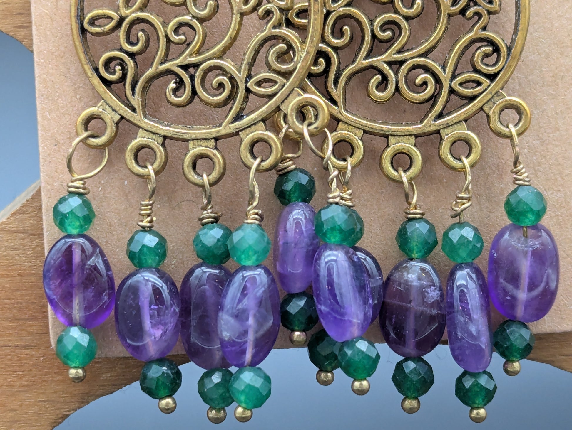 Close up of earrings with gold floral patterns set off by amethyst gemstones and green Swarovski crystal beads.