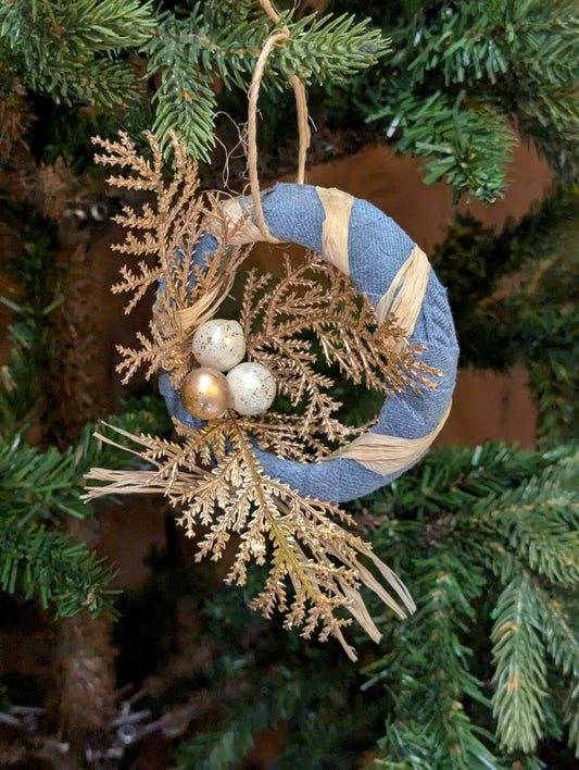 Decorative wreath ornament with blue flannel and raffia, gold and white berries, and faux gold cedar branches on a Christmas tree.