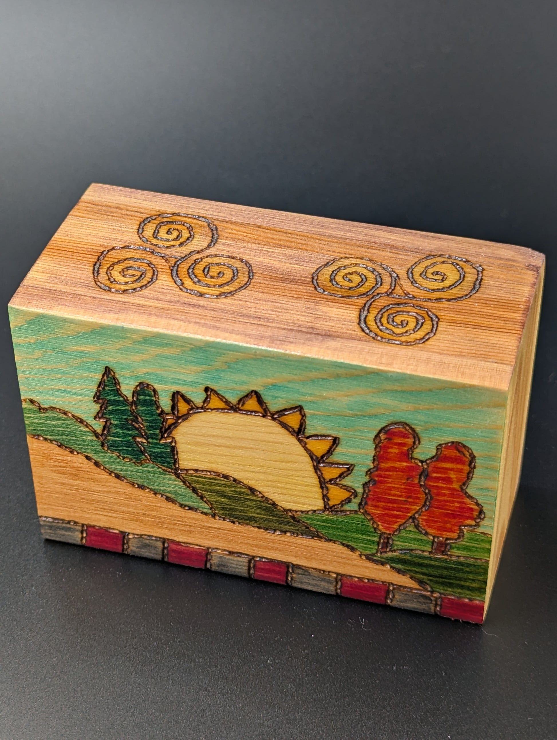 Wooden cube with artistic wood-burned and hand painted landscape of a sun, trees, and hills.